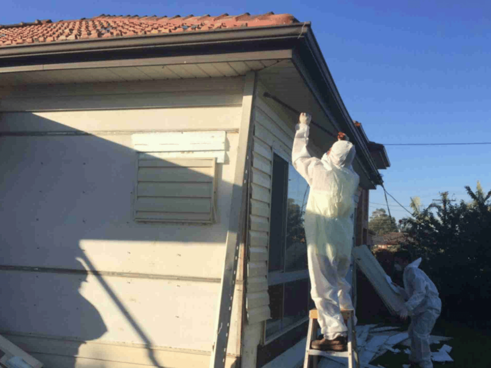 Two people in protective suits work on the exterior of a house; one stands on a ladder reaching the roof, while the other works near the ground. The house appears to be under renovation or removal of hazardous material.