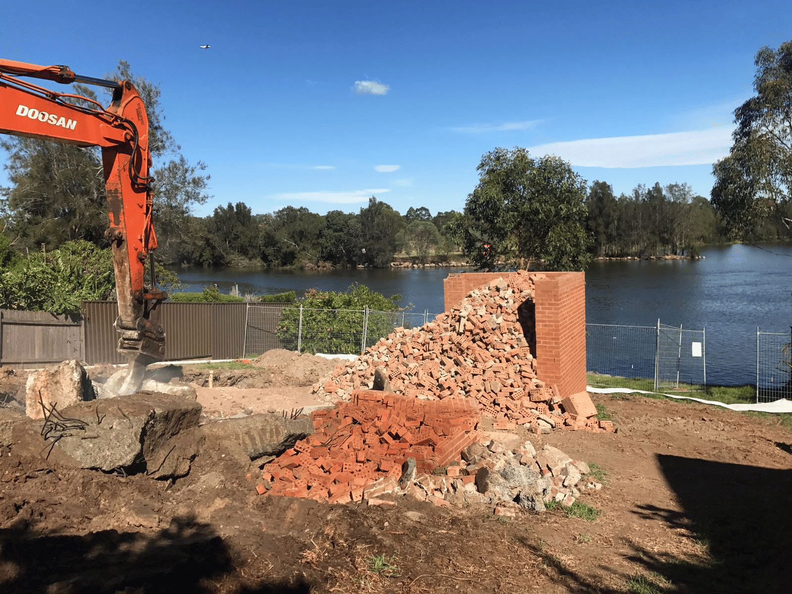 An orange excavator works near a partially demolished brick structure by a river, with piles of bricks and debris scattered on the ground. Trees and a clear blue sky are in the background.