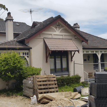 A house with a tiled roof and a small front porch is under renovation. Construction materials, wooden pallets, and sand are piled in the yard, with a metal fence partially surrounding the area.