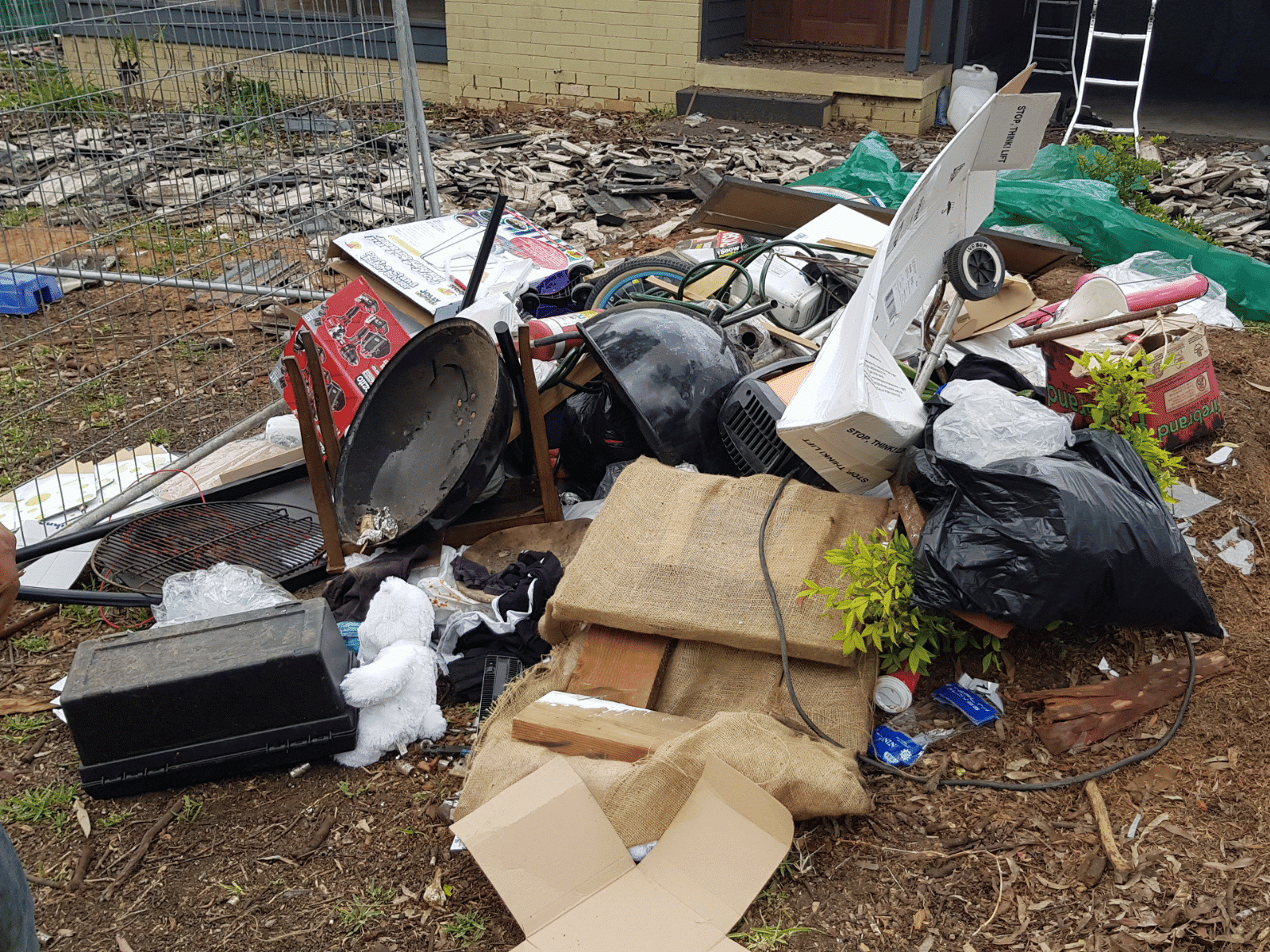 A large pile of assorted household junk and debris, including bags, cardboard boxes, a broken chair, and metal scraps, sits on bare ground in front of a house under renovation.