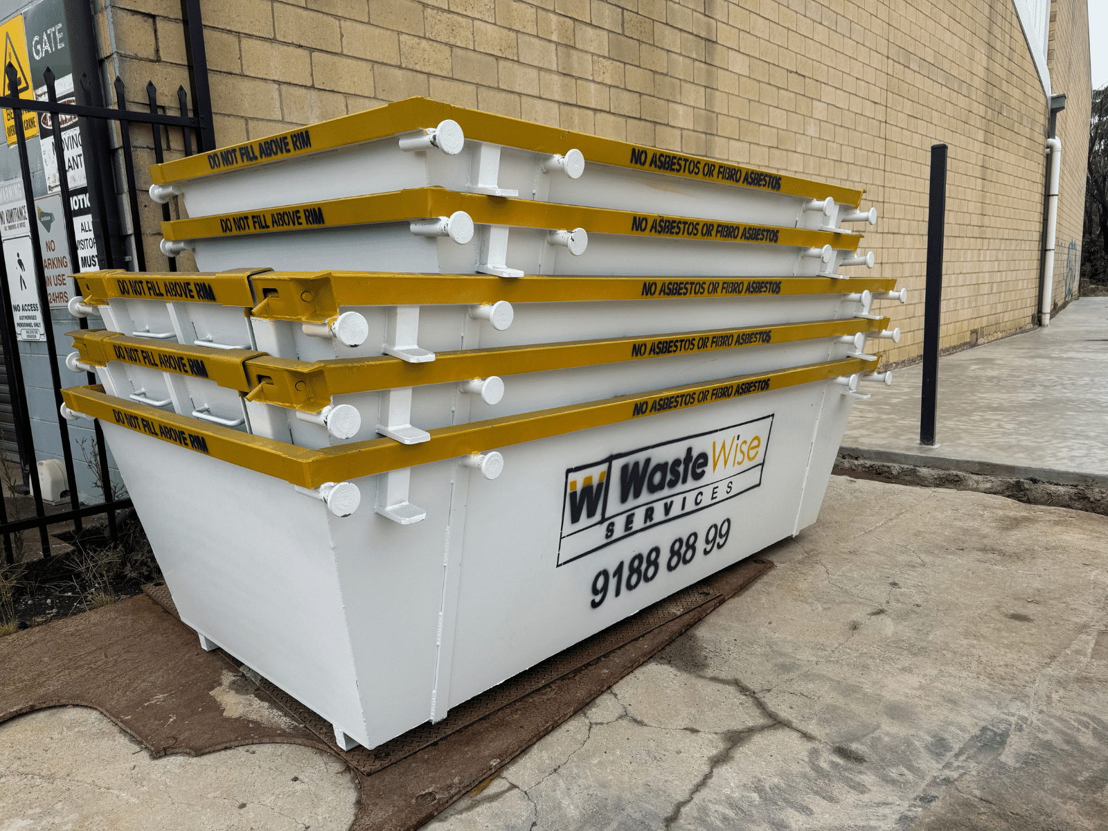 A stack of white metal waste containers with yellow rims, labeled "Waste Wise Services" and a phone number, placed on a concrete surface next to a brick wall.