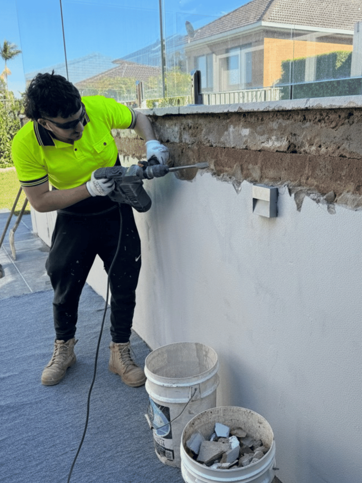 A man in a neon yellow shirt and black pants uses a power tool to remove bricks from an outdoor wall. Bits of debris fall into two white buckets below him, and a house is visible in the background.