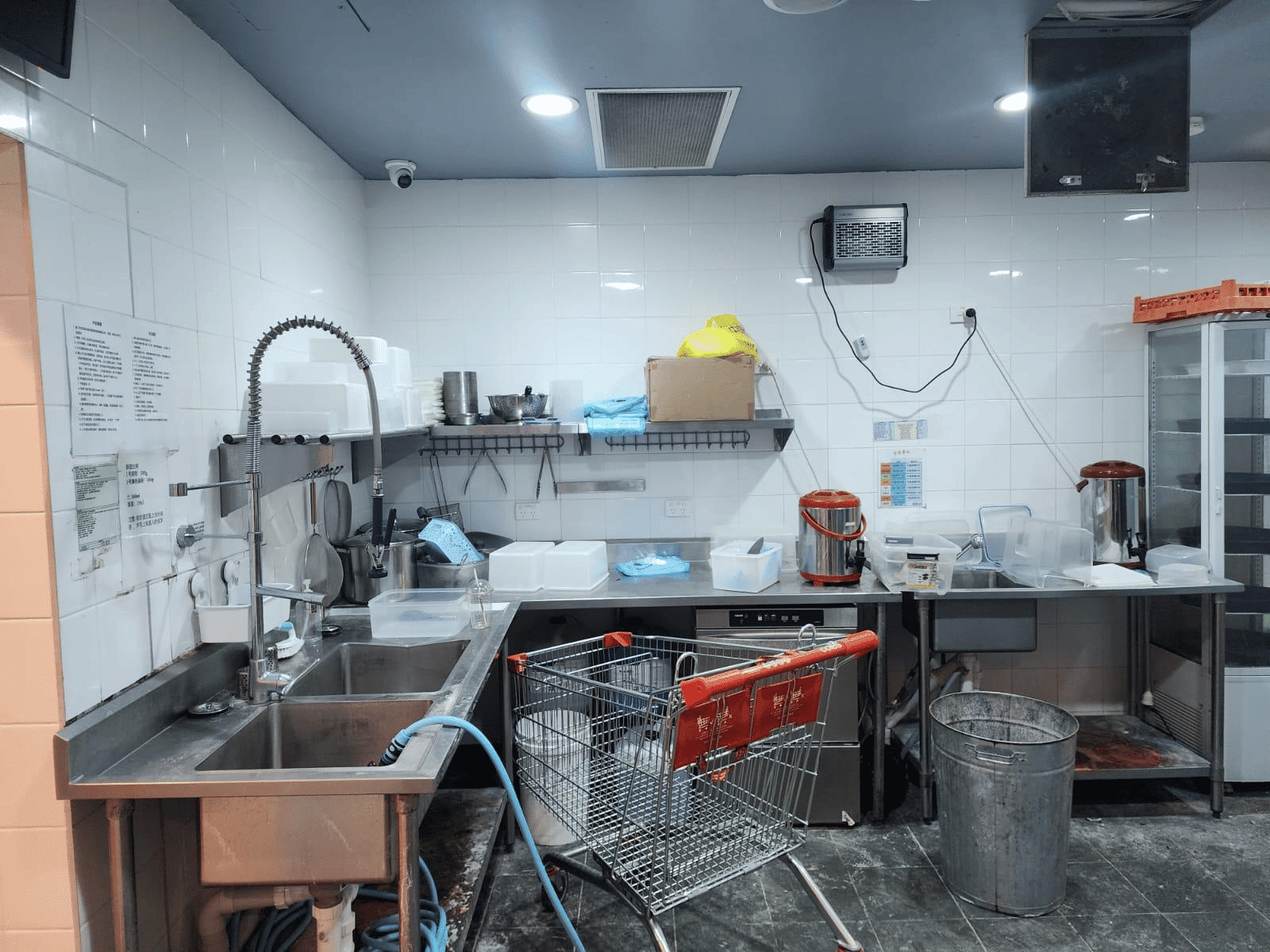 A commercial kitchen with a double sink, cleaning supplies, a shopping cart, plastic containers, metal shelves, a trash can, and a dishwasher. The floor is dark, and the walls are lined with white tiles.