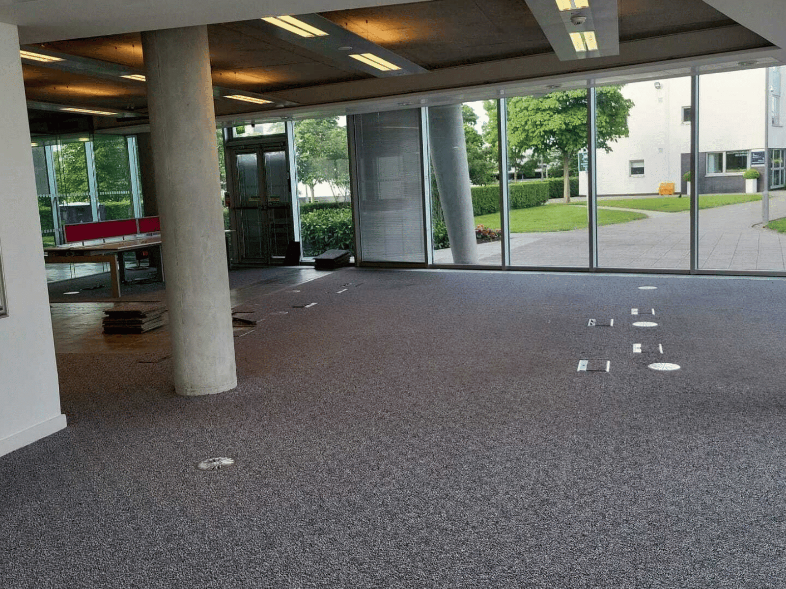 An empty office space with gray carpet, large floor-to-ceiling windows, and a concrete pillar. There are electrical outlets in the floor and a stack of carpet tiles near the entrance. Trees and buildings are visible outside.