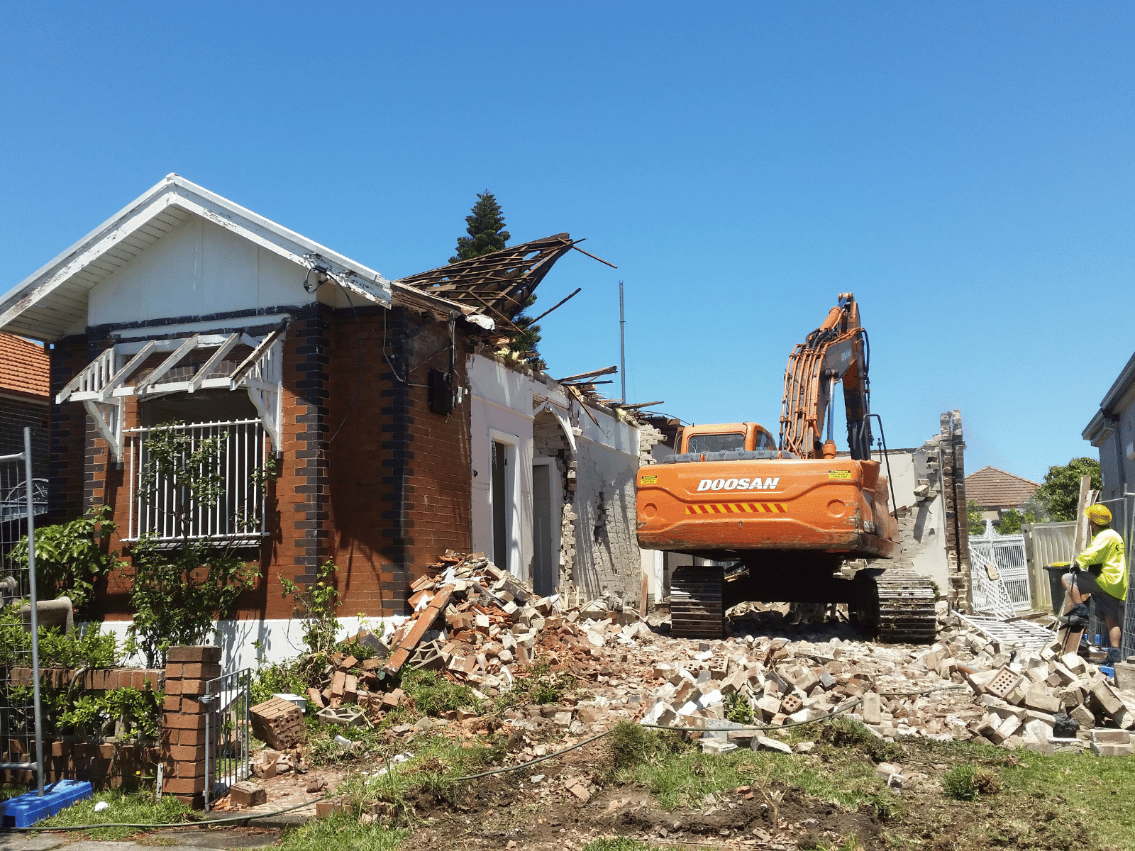 An orange excavator demolishes a brick house, with debris scattered around. A worker in a yellow vest and hard hat stands nearby under a clear blue sky.