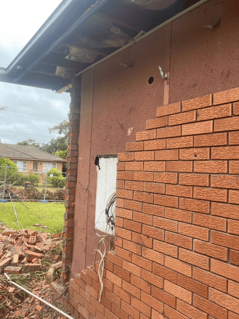 A partially demolished brick wall reveals exposed wiring and insulation in a house under renovation. Loose bricks and debris are scattered on the ground beside the wall. A grassy yard and suburban houses are visible in the background.