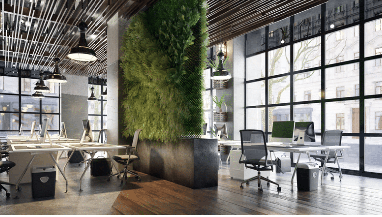 Modern open-plan office with large windows, several desks and chairs, indoor plants, and a vertical green wall. Natural light fills the space, and the décor features both wood and industrial elements.
