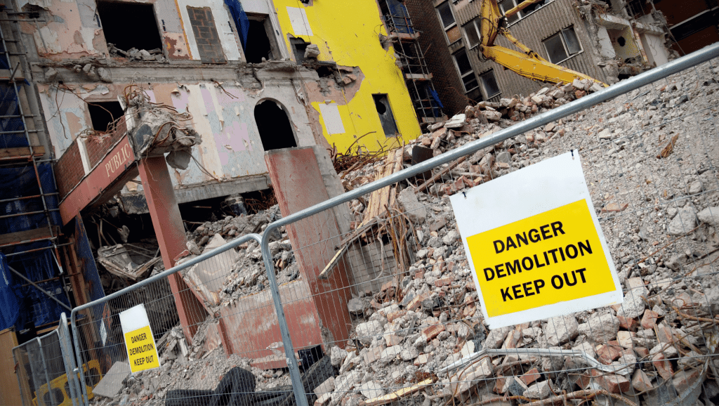 A partially demolished building surrounded by rubble and fencing. Yellow warning signs on the fence read "DANGER DEMOLITION KEEP OUT." Construction equipment is visible near the exposed structure.