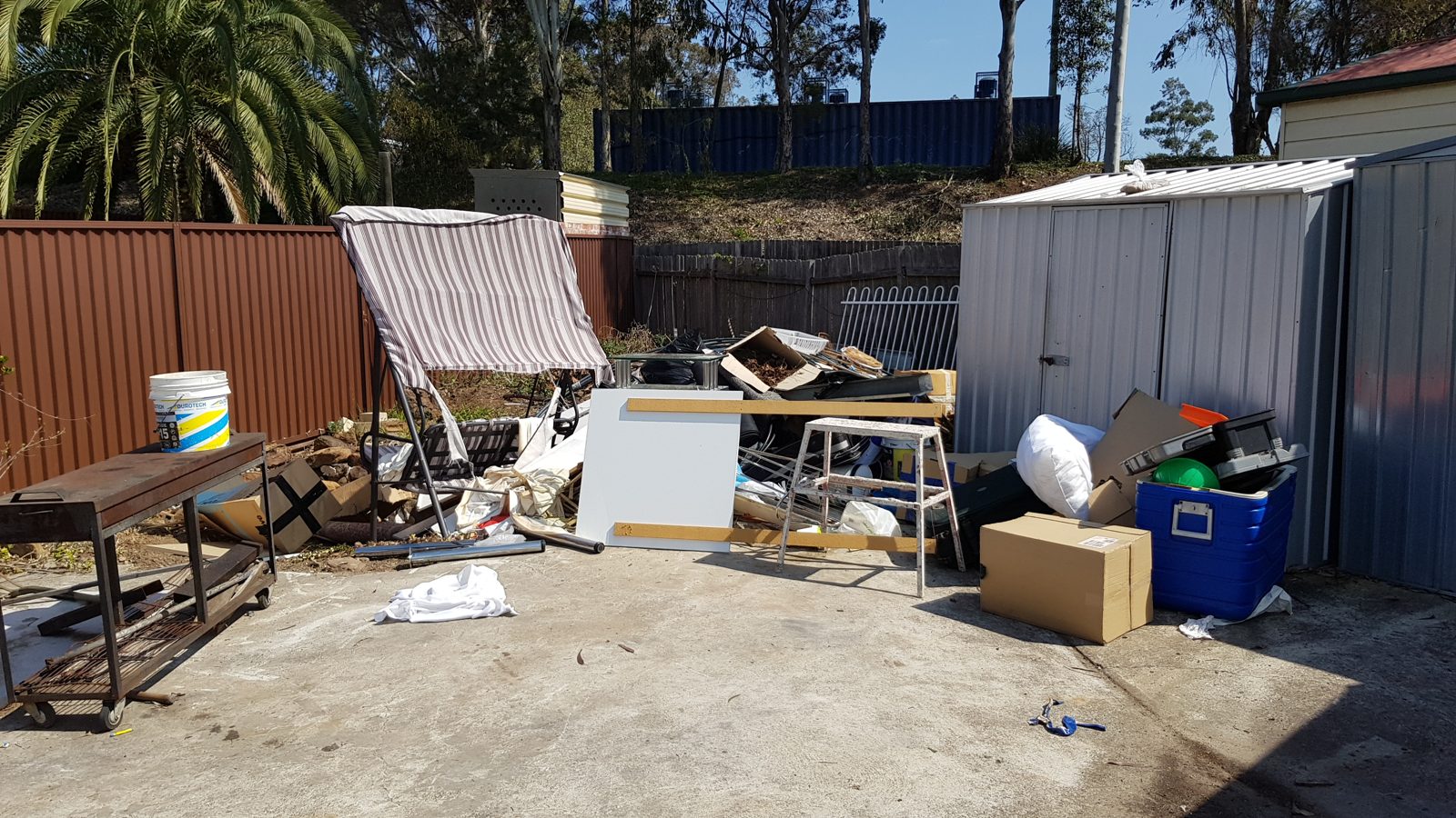 Outdoor area with various clutter, including cardboard boxes, a broken swing seat, a grill, a cooler, and scattered household items near two sheds on a concrete driveway. Trees and a fence are in the background.