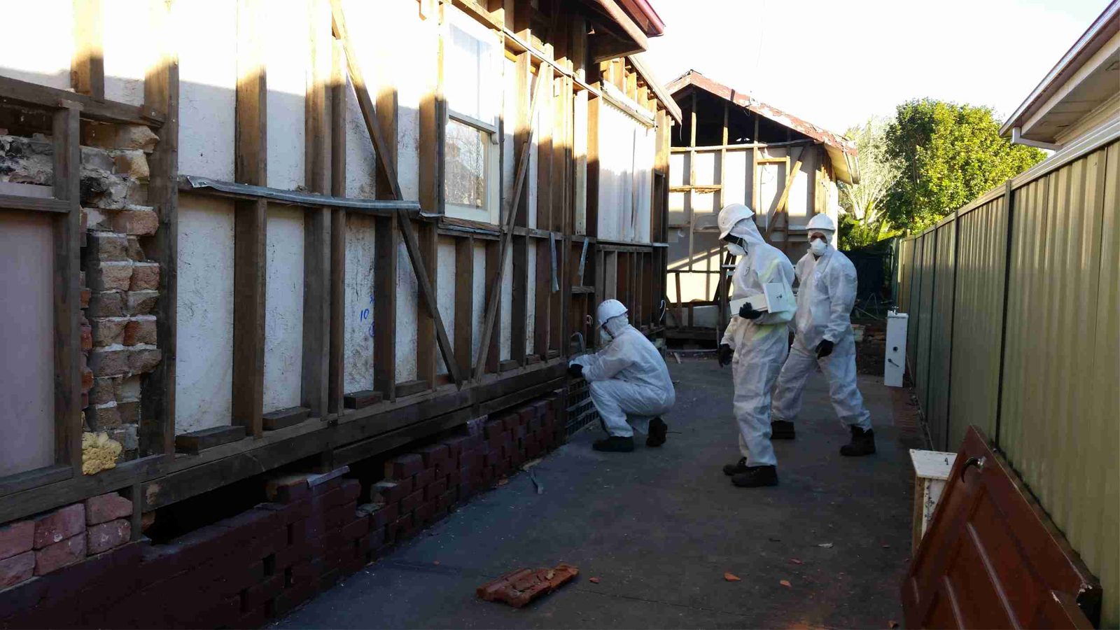 Three people in white protective suits work on the exposed wooden frame of a house, possibly during renovation or hazardous material removal, in an outdoor area between two buildings.