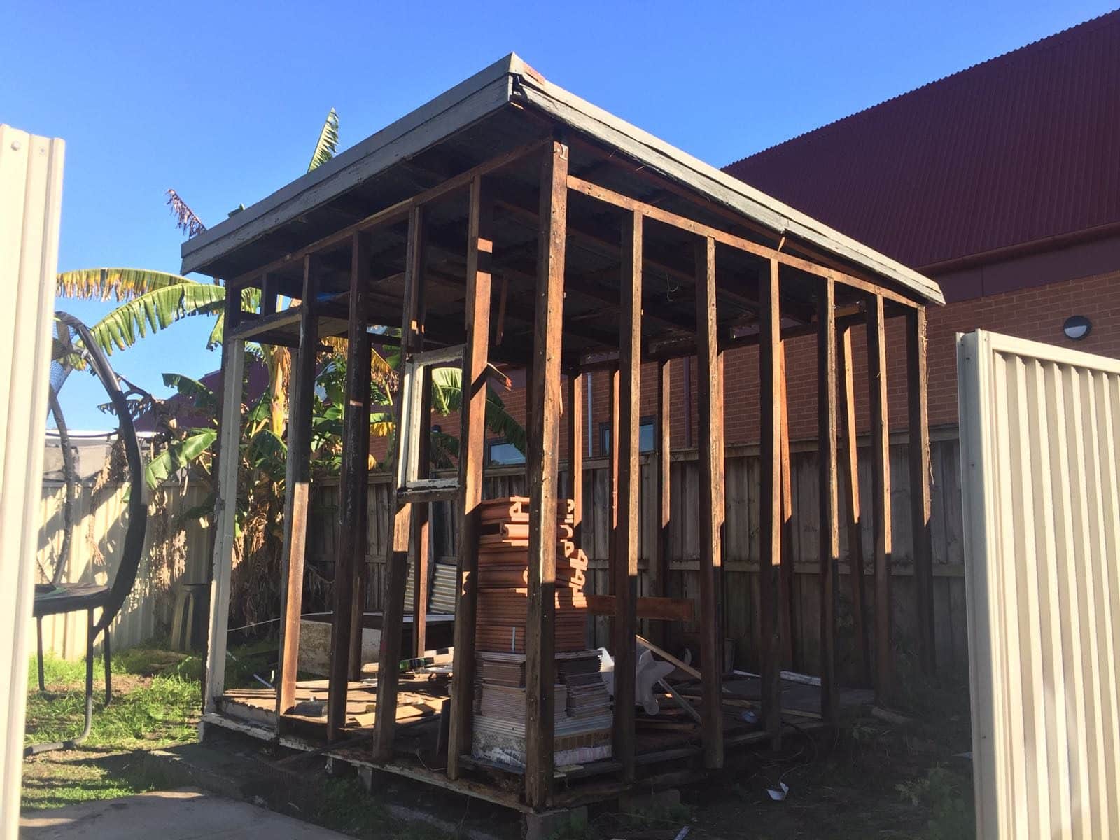 A partially constructed wooden shed stands in a yard, with an open frame and no walls. Construction materials and stacked items are inside, surrounded by a fence, banana plant, and brick building under a clear blue sky.