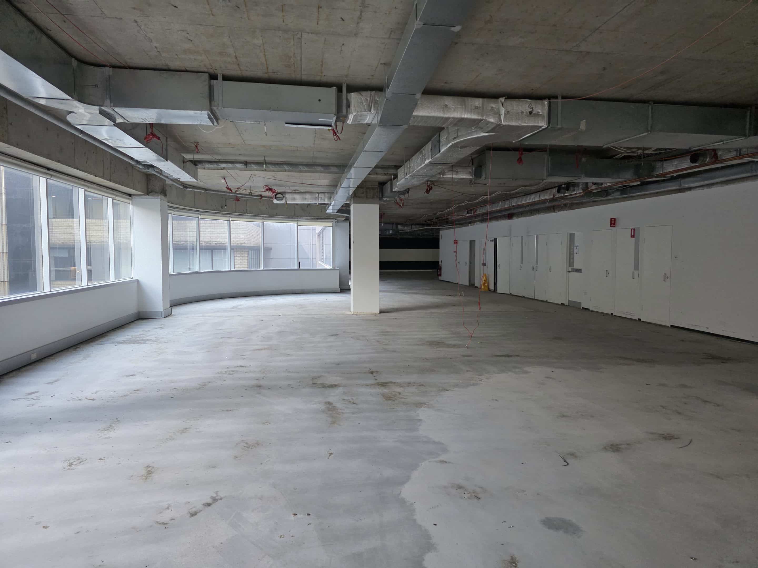 An unfinished office space with exposed concrete ceilings, ventilation ducts, large windows, and a row of white doors along the wall.