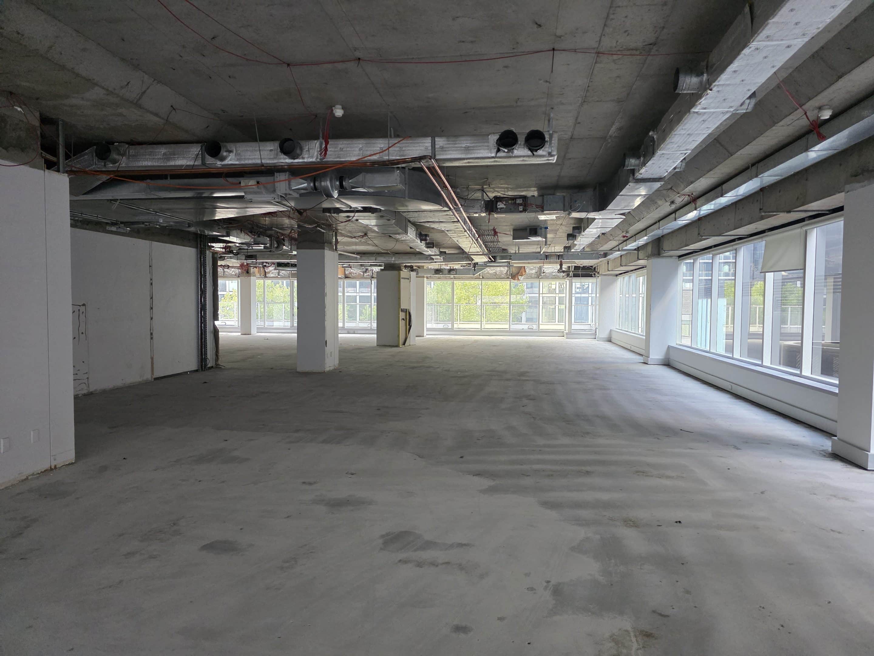 Large, empty commercial space under construction with exposed concrete ceilings, ventilation ducts, and floor-to-ceiling windows.