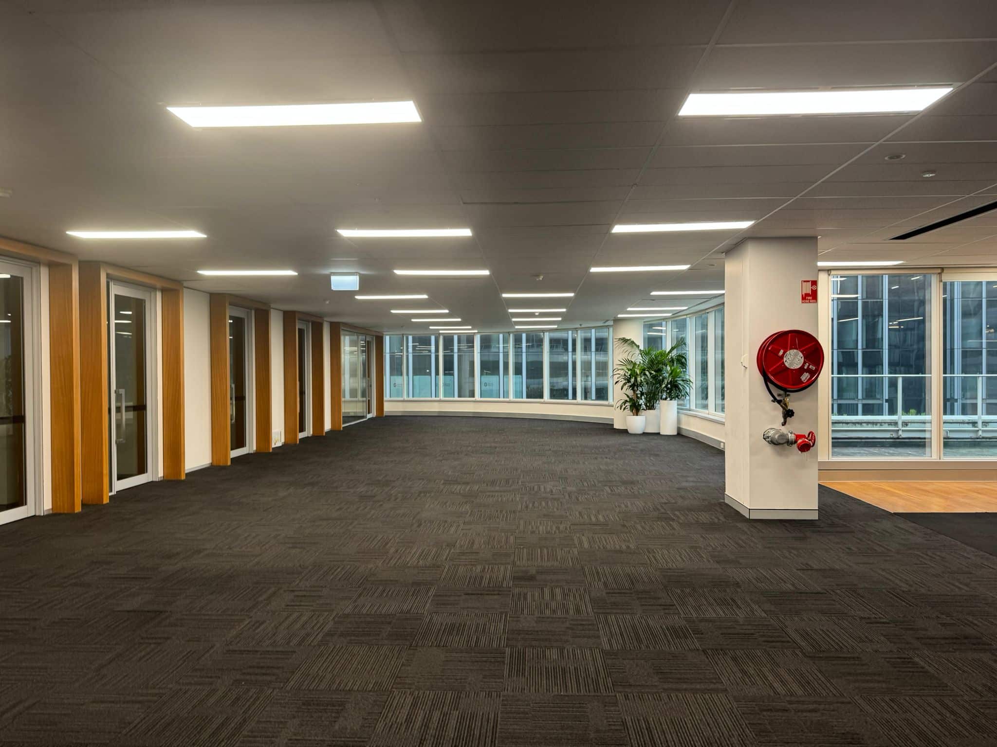 An empty office space with dark carpet, floor-to-ceiling windows, and a red fire hose reel mounted on a structural column.